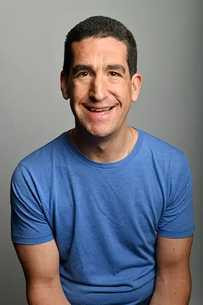 Darren Altman in a blue t-shirt smiling at the camera, arms by his side.